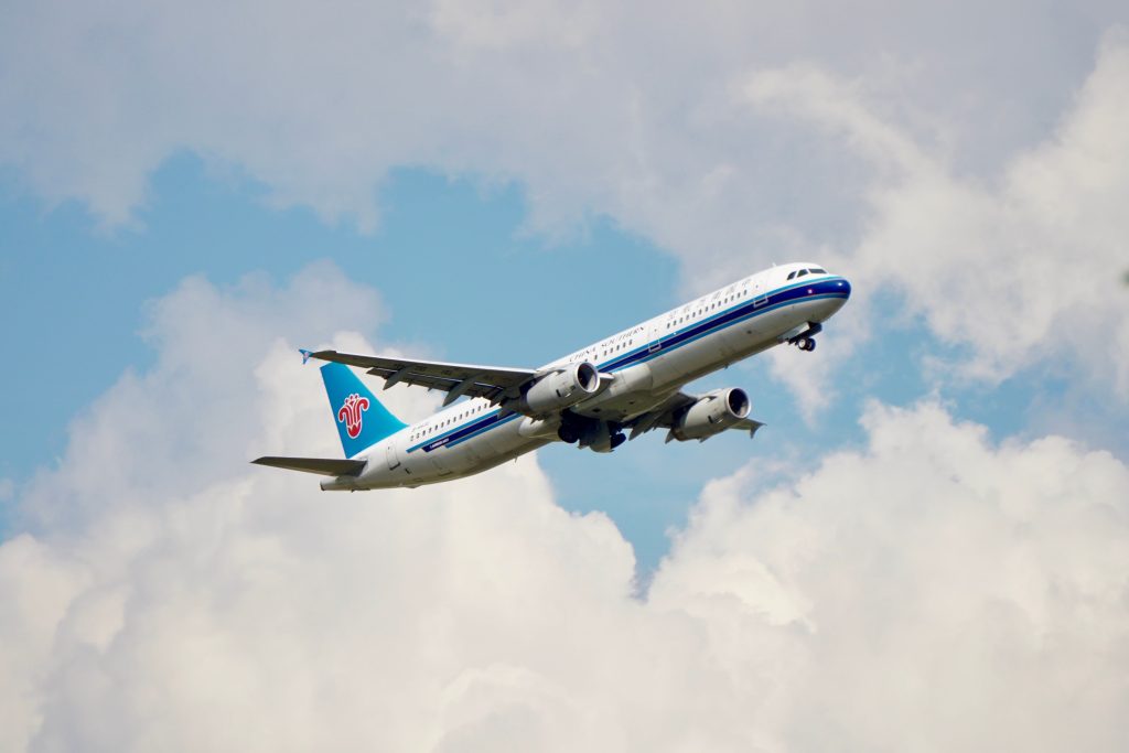 A commercial jet soaring in the broad daylight sky, surrounded by clouds.